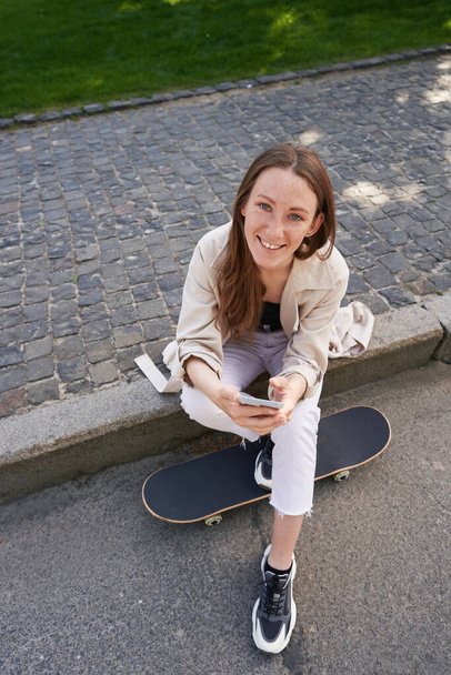 Mujer feliz sentada en asfalto junto al monopatín mientras sostiene el teléfono inteligente - Foto, Imagen