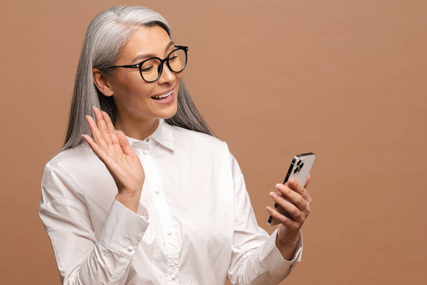 Carefree mature gray-haired lady has pleased virtual meeting, modern grandmother waving with hand. Glad senior woman is using smartphone for video connection isolated on beige - Fotó, kép
