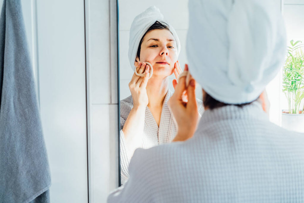 Woman in bathrobe with a towel on head looking in mirror, exfoliating facial skin with loofah sponge, removing makeup or doing cleansing routine, skincare treatment concept. Home Beauty self-care - Foto, Bild