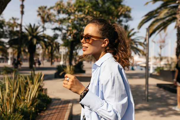 Stylish independent woman with collected hair wearing sunglasses and blue shirt is looking aside and walking in sunny street with exotic plants  - Photo, image