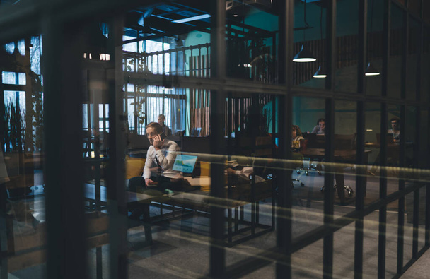 View through glass of people dressed in formal clothes sitting in spacious contemporary workspace and working on laptops and talking via smartphone - Fotoğraf, Görsel