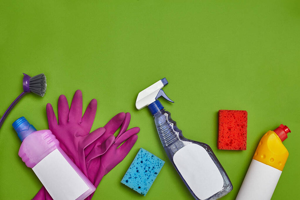 Ammattimaiset pesuaineet ja siivoustarvikkeet, kuten: värikkäät sienet, desinfiointiaine, spray ja puhdistusaineet astioille, joissa on tyhjä etiketti, violetti harja, violetti kumikäsineet vihreällä taustalla - Valokuva, kuva