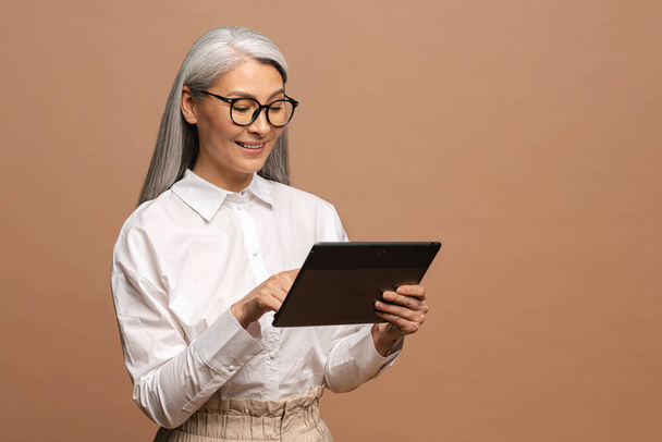 Modern elderly senior woman in formal wear using digital tablet isolated on beige. Portrait of mature female business person using online technology, computer app - Фото, изображение