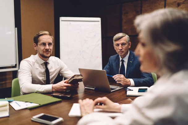 Group of wistful coworkers sitting at table in meeting room with laptop and surfing internet while discussing details of business plan - Photo, image