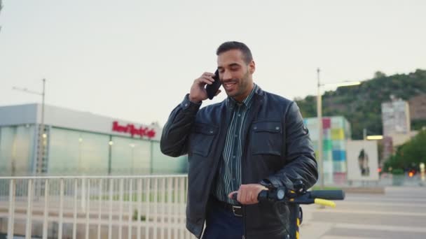 Jovem homem elegante conversando com amigo no smartphone enquanto caminha pela rua com e-scooter. cara atraente usando scooter elétrico ao ar livre - Filmagem, Vídeo