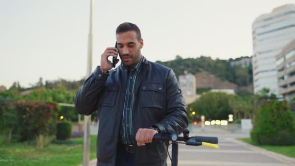 Aantrekkelijke man in leren jasje pratend over smartphone terwijl hij over straat loopt met een e-scooter. Jongeman gaat door de stad met elektrische scooter - Video