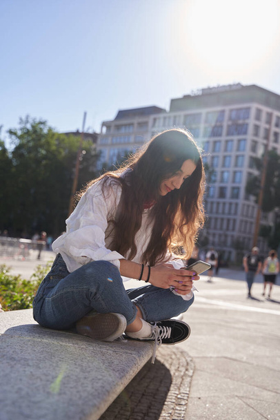 Улыбающаяся молодая кавказская хипстерша с длинными волосами, держащая смартфон, используя мобильное приложение, сидящее снаружи. Вертикальный вид - Фото, изображение
