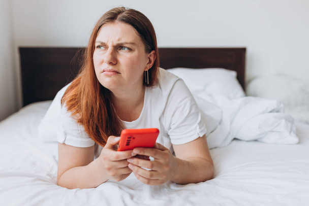 Zdenerwowana młoda kobieta leżąca w łóżku czuje się chory lub zaniepokojony za pomocą nowoczesnego gadżetu komórkowego, smutne tysiąclecia kobieta relaks w sypialni hold smartphone czytać złe wieści - Zdjęcie, obraz