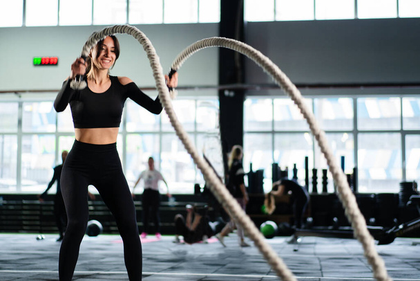 Powerful woman training battle ropes at cardio workout in dark gym. Professional athlete exercise fitness sport club equipment. Strong bodybuilder lifting weights. Athletic person effort - Fotó, kép