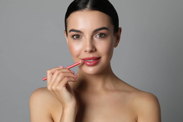 Pretty young woman applying beautiful pink lip pencil on grey background - Foto, imagen