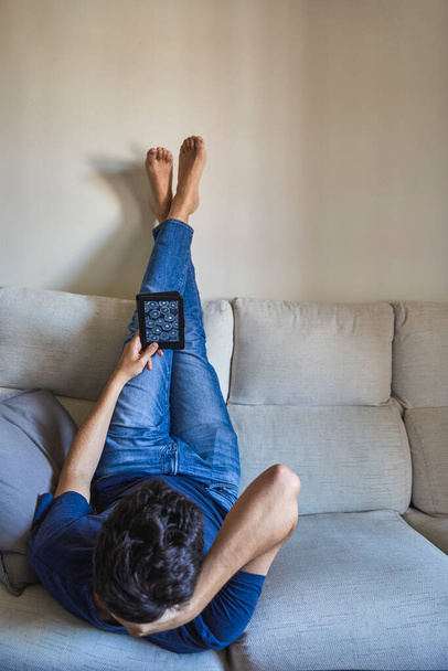 Молодой человек лежит с ногами на стене лицом вниз и читает электронную книгу на диване - Фото, изображение