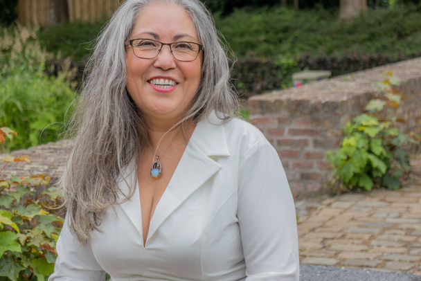 Portrait of mature woman with happy smiling expression, attentively looking at camera, straight gray hair, tanned skin, glasses, light makeup, relaxed day in garden, green vegetation in background - Photo, Image