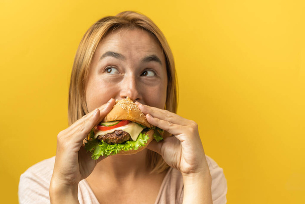 Ritratto di bella giovane donna caucasica con capelli biondi che si gode un hamburger su uno sfondo giallo. Concetto di dieta - Foto, immagini