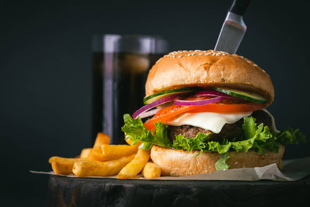 Big burger Patty wołowiny z serem pomidor fioletowy ogórek cebula i sałata obok frytki na drewnianym czarnym stoisku na czarnym tle - Zdjęcie, obraz