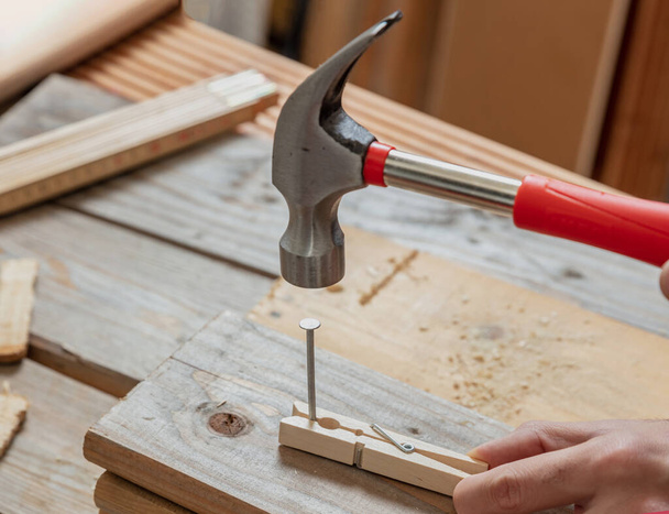 Male hand hold hammer and nail a clothespin, wooden work bench table background. DIY, home repair and fix. - Photo, Image