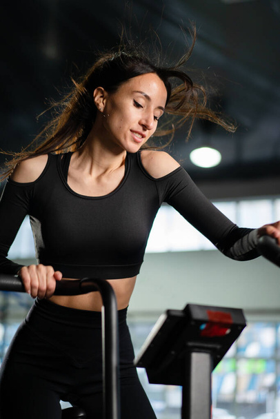 Young Athletic middle aged woman training on air resistance bike, cross training workout set in gym. Active woman spinning a air bike in gym with trainers. female training on air bike. - Photo, Image