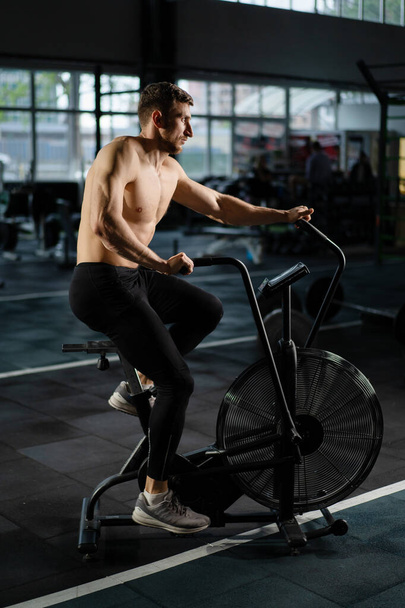Young Athletic middle aged man training on air resistance bike, cross training workout set in gym. Active man spinning a air bike in gym with trainers. Male training on air bike. - Fotoğraf, Görsel