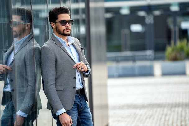 Outdoor photo of handsome elegant man wearing fashionable clothes and sunglasses, posing on the wall with reflections. A lot of copy space. City stale. Male model. - Φωτογραφία, εικόνα