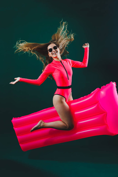 Atractiva mujer alegre en un traje de baño rosa, sobre un fondo aislado tiene un colchón inflable entre sus piernas. Concepto de vacaciones. Chica delgada en gafas de sol en un colchón de aire - Foto, imagen