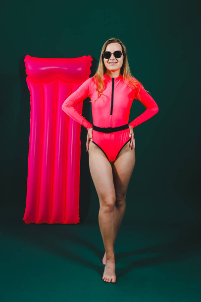 Atractiva mujer delgada en un traje de baño rosa, sobre un fondo aislado. Concepto de vacaciones, vacaciones de verano. Chica delgada en gafas de sol contra el fondo de un colchón inflable de color rosa - Foto, imagen