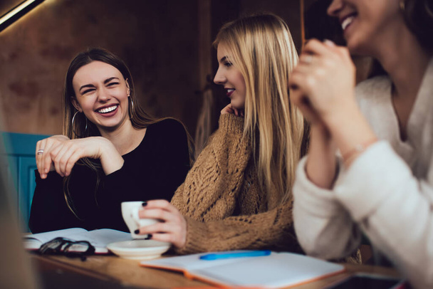 Young beautiful women in casual clothing laughing while spending time together and working on collaborative project in cozy time cafe - Photo, Image