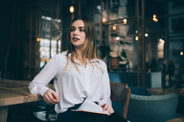 Young lady in trendy clothes sitting near wooden table and flipping pages in notebook while resting in contemporary cafe and looking dreamily away - Foto, Imagen