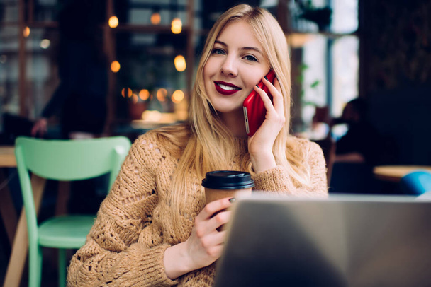 Positive female freelancer in casual wear and red lipstick resting in modern cafe with laptop and to go coffee while looking at camera - Photo, Image