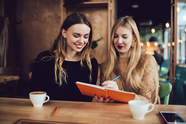 Positive female friends in casual clothes sitting at wooden table and writing ideas in copybook while resting together in cafe - Foto, imagen