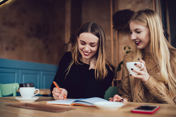 Happy female friends in casual wear sitting at wooden table and taking notes in notebook while resting together in cafe - Фото, зображення