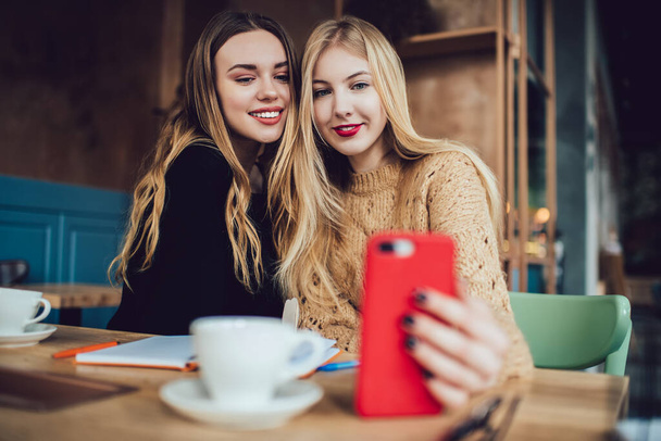 Smiling female friends in casual wear sitting at wooden table in cafe and taking selfie on smartphone while chilling together - Foto, Imagem