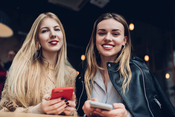From below of cheerful young female friends sitting in cafe and holding mobile phones in hands while looking at camera - Foto, Bild