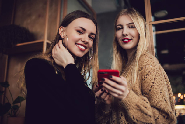 Low angle of positive female friends in casual wear smiling and messaging on smartphone while resting in modern cafe while spending time together - 写真・画像