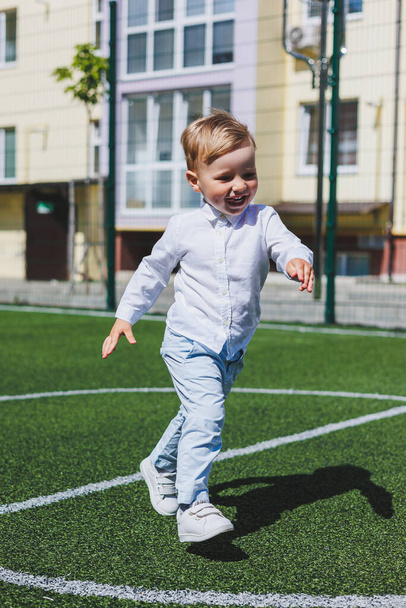 Ένα μικρό αγόρι 2 ετών με πουκάμισο και παντελόνι τρέχει και γελάει. Ένα παιδί σε μια ηλιόλουστη μέρα χαίρεται και χαμογελάει. - Φωτογραφία, εικόνα