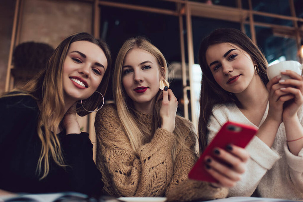 Θετικές νεαρές όμορφες γυναίκες σε casual ρούχα κοιτάζοντας κάμερα, ενώ έχουν ξεκουραστεί και να περνούν το χρόνο τους μαζί σε ζεστό καφέ - Φωτογραφία, εικόνα