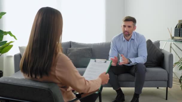 Emotional smiling adult man patient with mental health problem sitting on couch talking to therapist. Positive woman psychologist with clipboard listen client at psychotherapy session in clinic office - Footage, Video