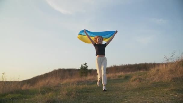 Młoda blondynka biegnie z ukraińską flagą w rękach nad głową na zachodzącym słońcu. Uśmiecha się. Znak zwycięstwa. Wysokiej jakości materiał filmowy FullHD - Materiał filmowy, wideo