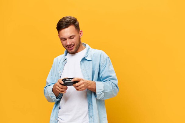 Cieszył się opalony przystojny gracz człowiek w niebieskim podstawowym t-shirt trzymać joystick spojrzeć na gamepad pozowanie izolowane na pomarańczowy żółty tło studio. Skopiuj Kosmiczny Baner Mockup. Koncepcja stylu życia ludzi - Zdjęcie, obraz