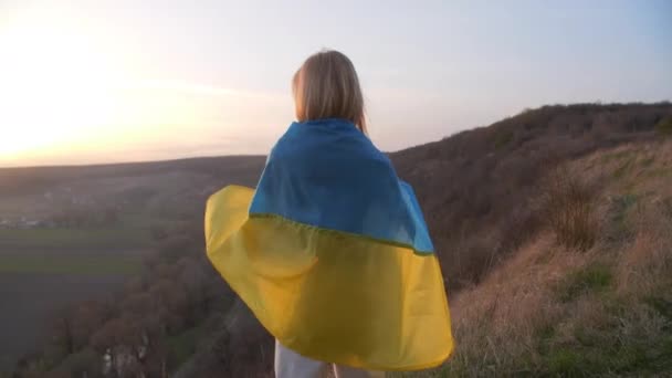 Une jolie femme blonde sourit et se promène avec le drapeau ukrainien. Elle regarde le coucher du soleil et se tient derrière avec le drapeau enveloppé. Images 4k de haute qualité - Séquence, vidéo