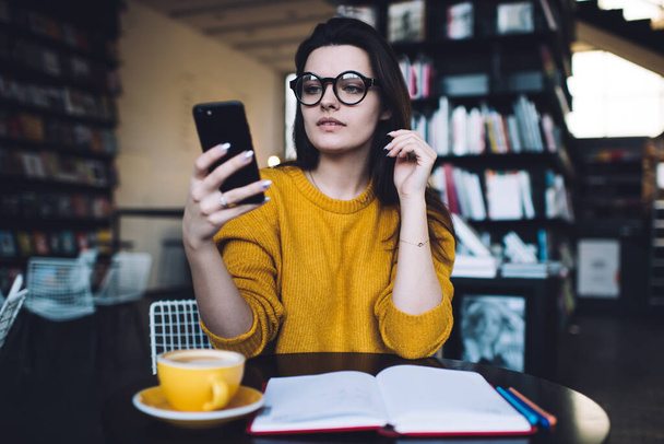 Concentrated female in eyeglasses sitting at black table near bookshelf and using cellphone while spending time alone in cozy cafeteria - 写真・画像