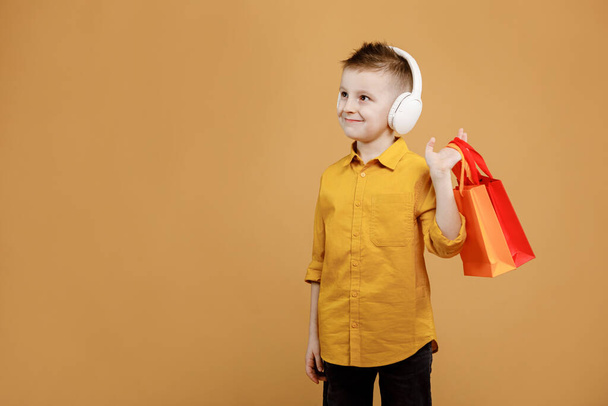 Image of smiling boy holding bags with presents or shoppings on yellow background. child in white headphone. Shopper with many colored paper bags. Holidays sales and discounts. Cyber monday. - Fotó, kép