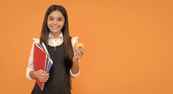 Χαρούμενο σχολικό χαμόγελο κρατώντας το μήλο και τα βιβλία πορτοκαλί φόντο αντίγραφο χώρο, σχολείο. - Φωτογραφία, εικόνα