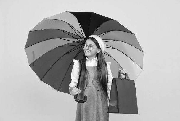 szezonális eladás. tinédzser gyerek színes napernyő alatt. Gyerek svájcisapka szivárvány esernyővel. Őszi szezon. esős időjárás előrejelzés. Vissza az iskolába. élénk esővédelem. boldog lány szemüvegben tartsa bevásárló táska. - Fotó, kép