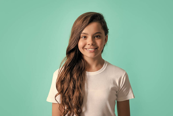 cheerful kid with curly hair. beauty and fashion. female fashion model. pretty look of young girl in white shirt. portrait of happy child on blue background. express positive emotions. - Fotografie, Obrázek