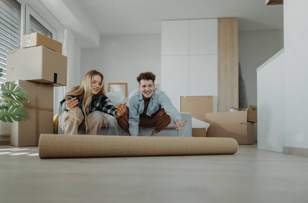 A cheerful young couple in their new apartment, rolling out carpet. Conception of moving. - Foto, immagini