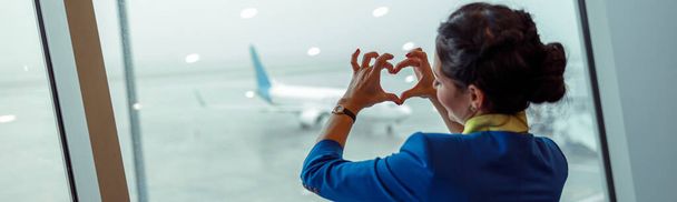 Assistente de bordo fêmea fazendo sinal de coração com as mãos enquanto está ao lado da janela e desfrutando de vista para o aeródromo - Foto, Imagem