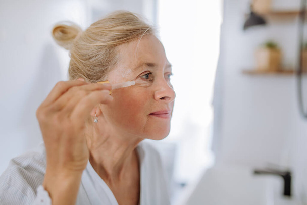 A beautiful senior woman in bathrobe, moisturizing skin by pipette with serum enjoying treatment procedure. - Фото, зображення