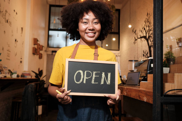 One African American female startup barista stands at casual cafe door, looks at camera, and shows open sign, happy and cheerful smiles with coffee shop service jobs, and new business entrepreneurs. - Photo, Image