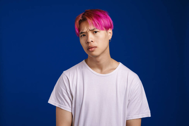 Ázsiai fiú rózsaszín haj ráncolás és nézi kamera elszigetelt felett kék háttér - Fotó, kép