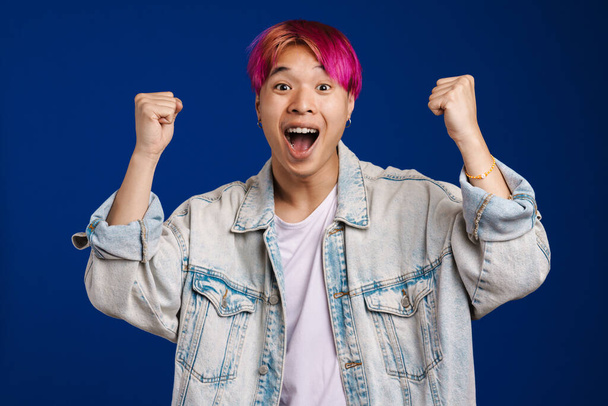 Asian boy with pink hair screaming while making winner gesture isolated over blue background - Fotografie, Obrázek