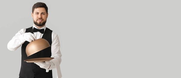 Schöner männlicher Kellner mit Tablett und Cloche auf hellem Hintergrund mit Platz für Text - Foto, Bild